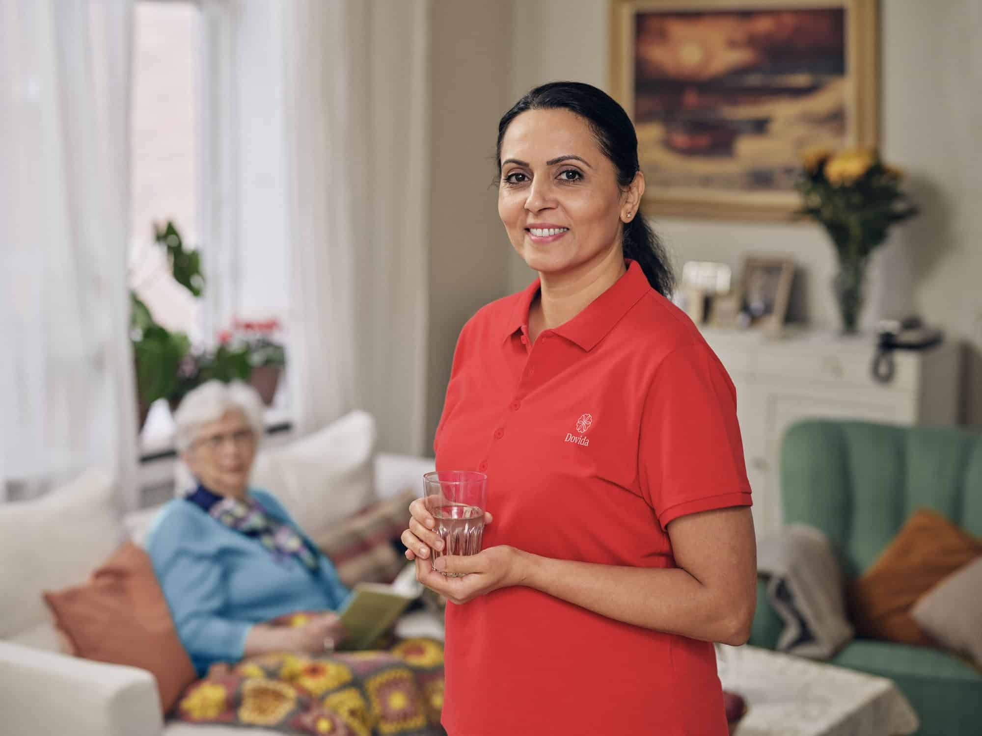 A caregiver in a red Dovida shirt smiles warmly while holding a glass, embodying care and compassion in Dovida careers