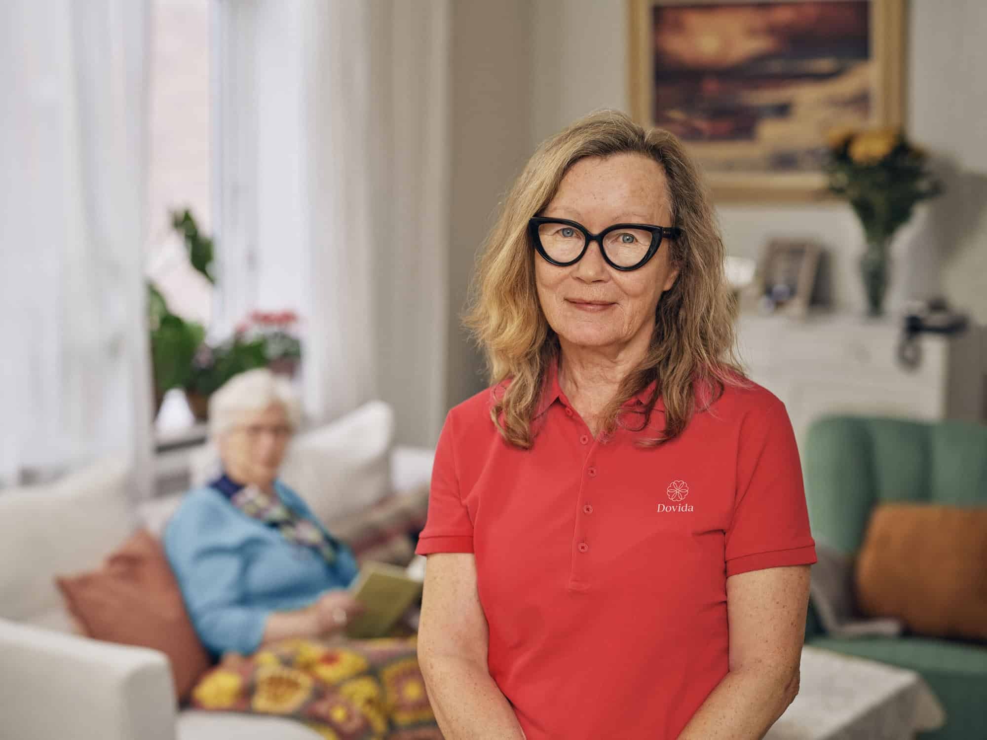 A confident woman in glasses and a red Dovida shirt smiles in her home care office, radiating professionalism.