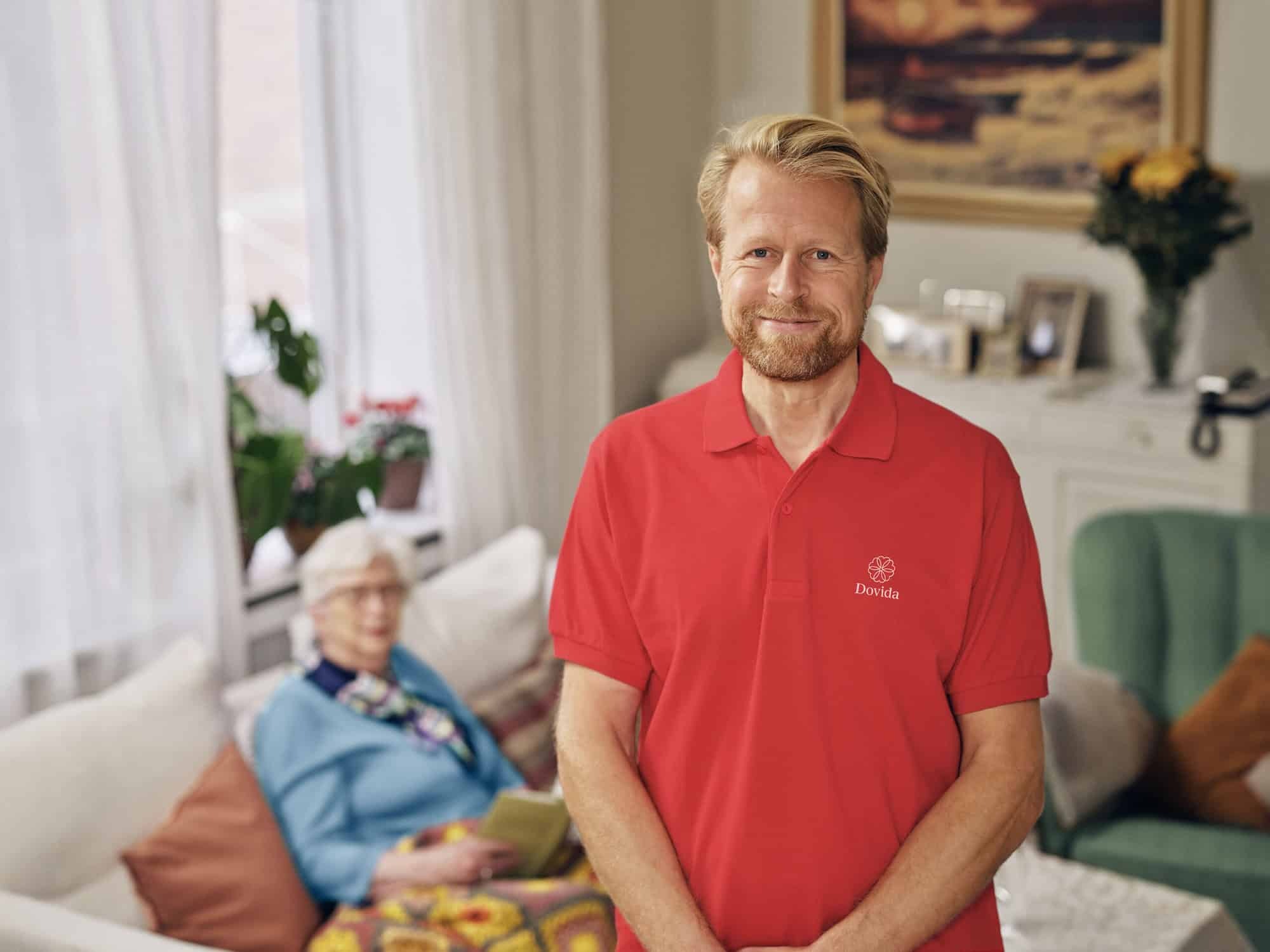 An in home caregiver in a red Dovida shirt stands confidently, ready to assist as a home caregiver for the elderly.